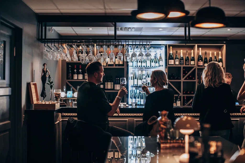 Gäster med vinglas vid bardisken i vinbaren, framför upplysta vinhyllor och hängande glas