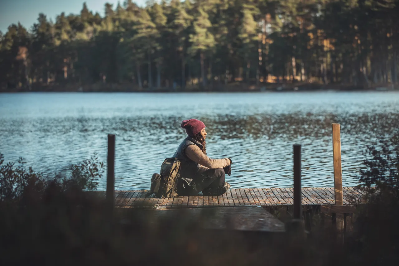 En person sitter på en brygga vid en skogssjö i kvällssol
