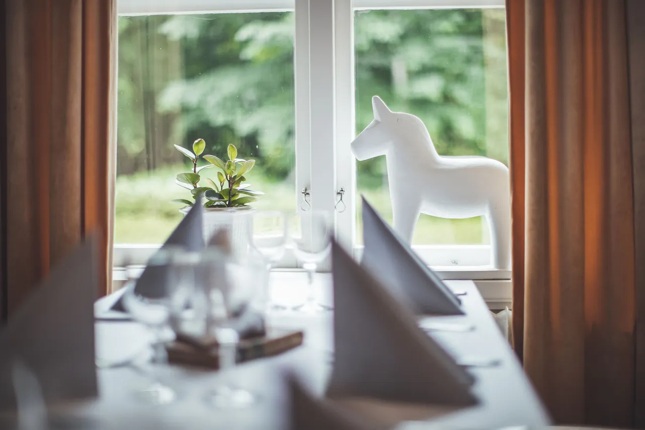 Dukat bord vid ett fönster mot grönskan, med en vit dalahäst i fönstret