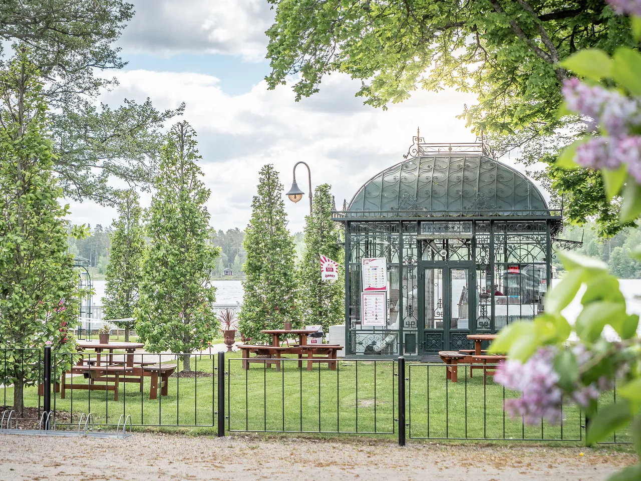 Glasscaféet i det gröna orangeriet vid sjön, med bänkbord på gräsmattan och syrener i förgrunden