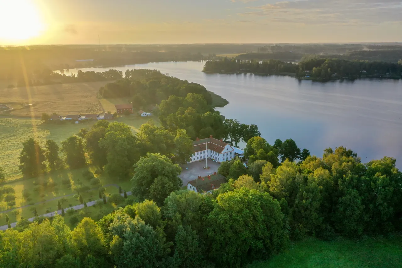 Herrgården bland lummiga lövträd vid en stor sjö, sedd från ovan i lågt morgonljus