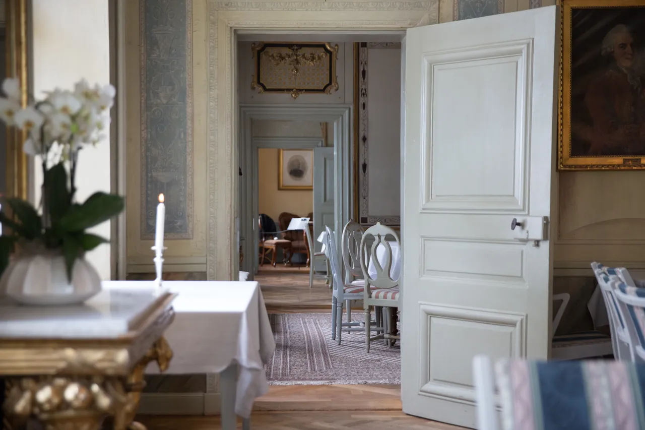 Vy genom en öppen dörr till en ljus salong med dukade bord, en orkidé och ett tänt ljus i förgrunden