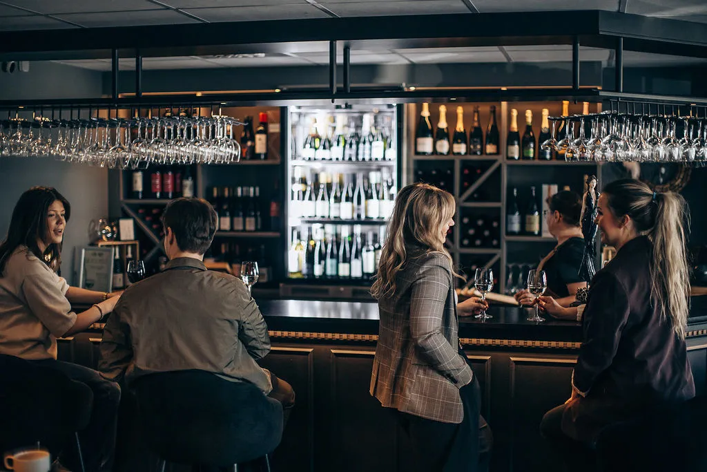Gäster i samspråk vid bardisken i vinbaren, med vinhyllor och hängande glas i varmt ljus