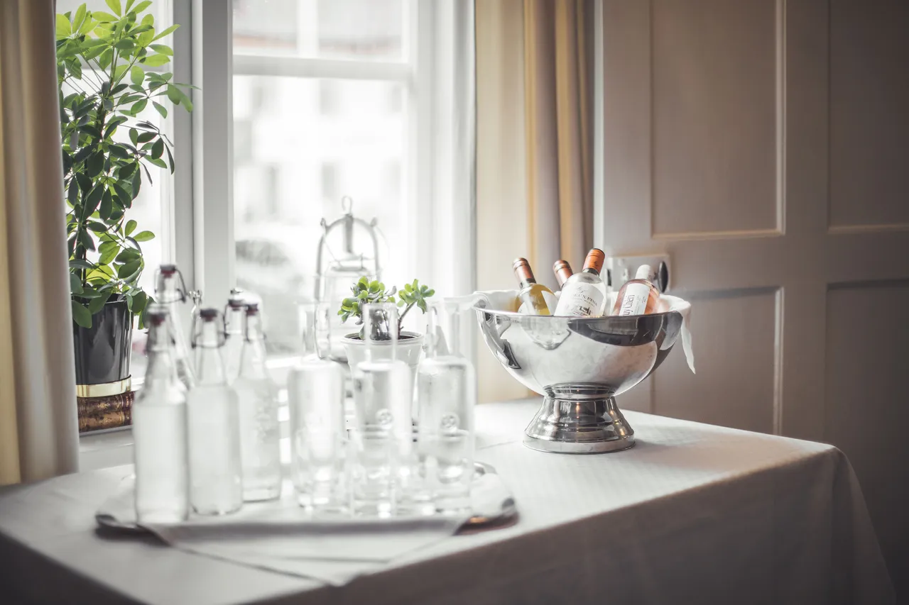 Glaskaraffer, glas och en silverkylare med flaskor på ett dukat bord vid fönstret
