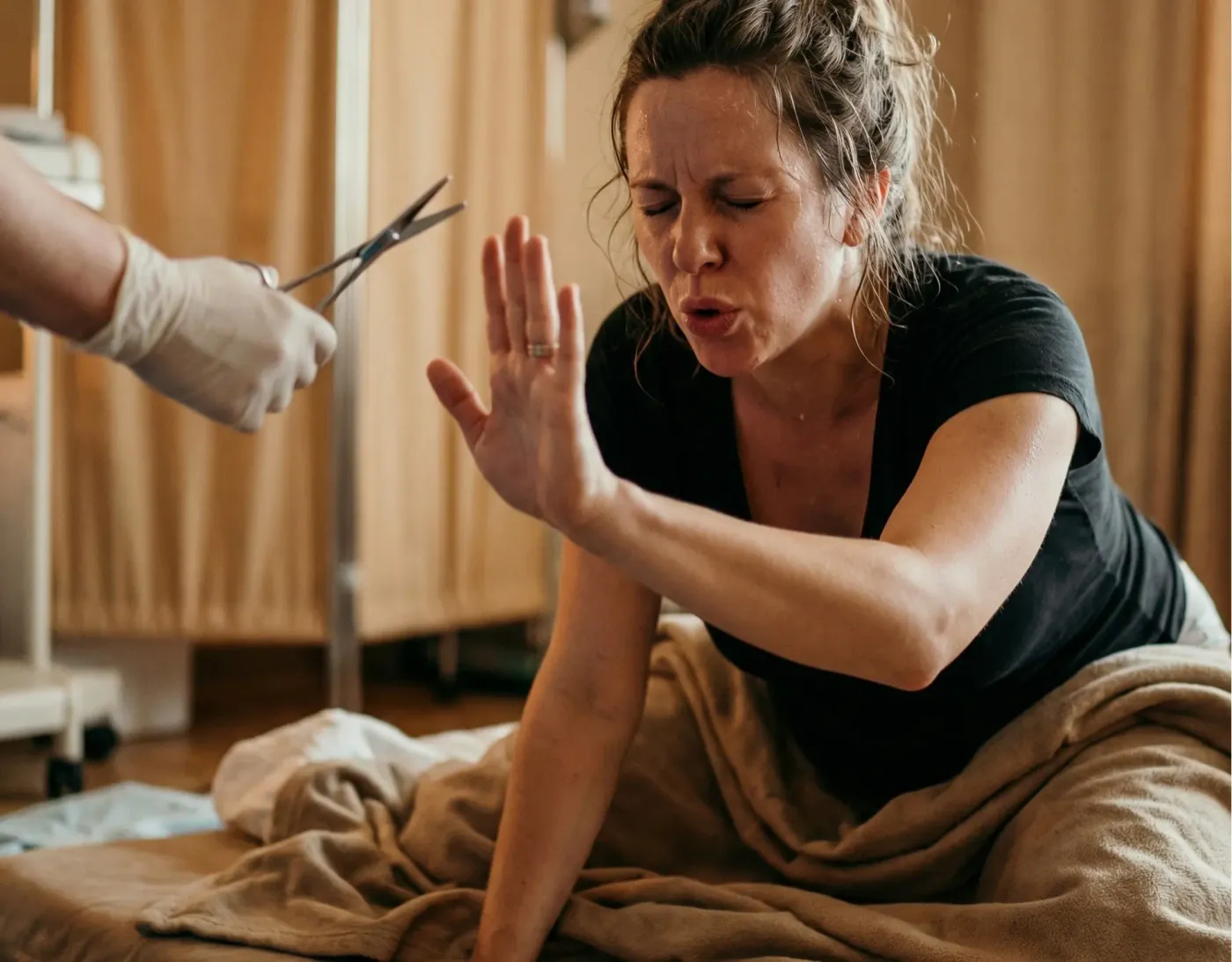 Une femme en plein travail, les yeux fermés, lève la main pour arrêter une main gantée qui tient des ciseaux