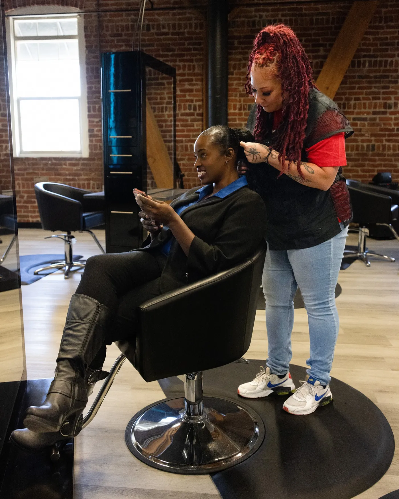 A Style City stylist styles a smiling client by the brick wall and big window, empty stations behind them