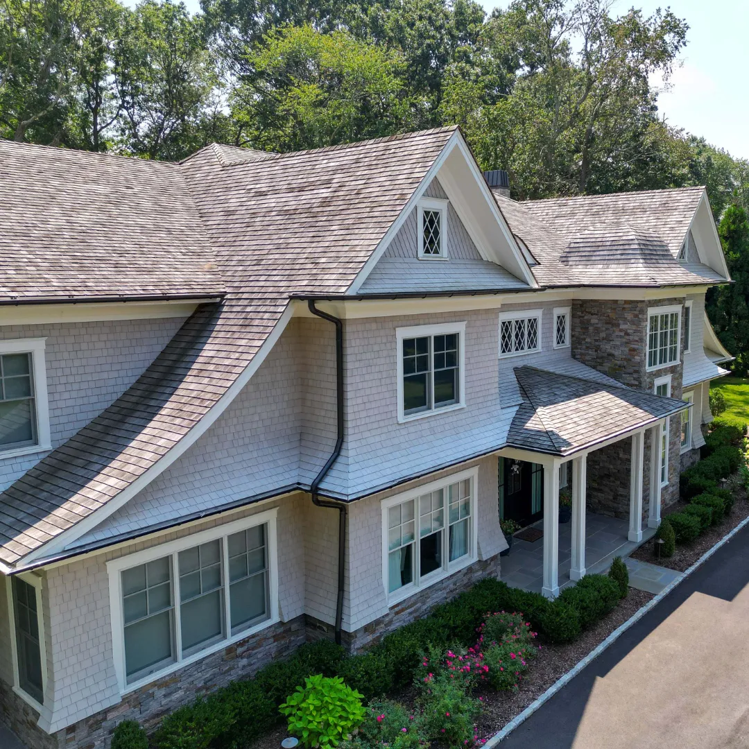 A shingle-style Prestige home seen from above: gabled dormers, a stone base and a covered front porch among mature trees