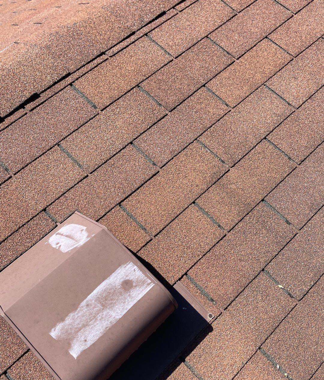 Brown metal roof vent with chalk marks on dents, surrounded by brown asphalt shingles