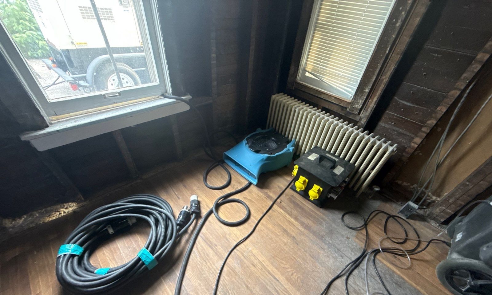 Air mover and dehumidifier hoses on a wood floor in a room stripped to lath beside a window and radiator