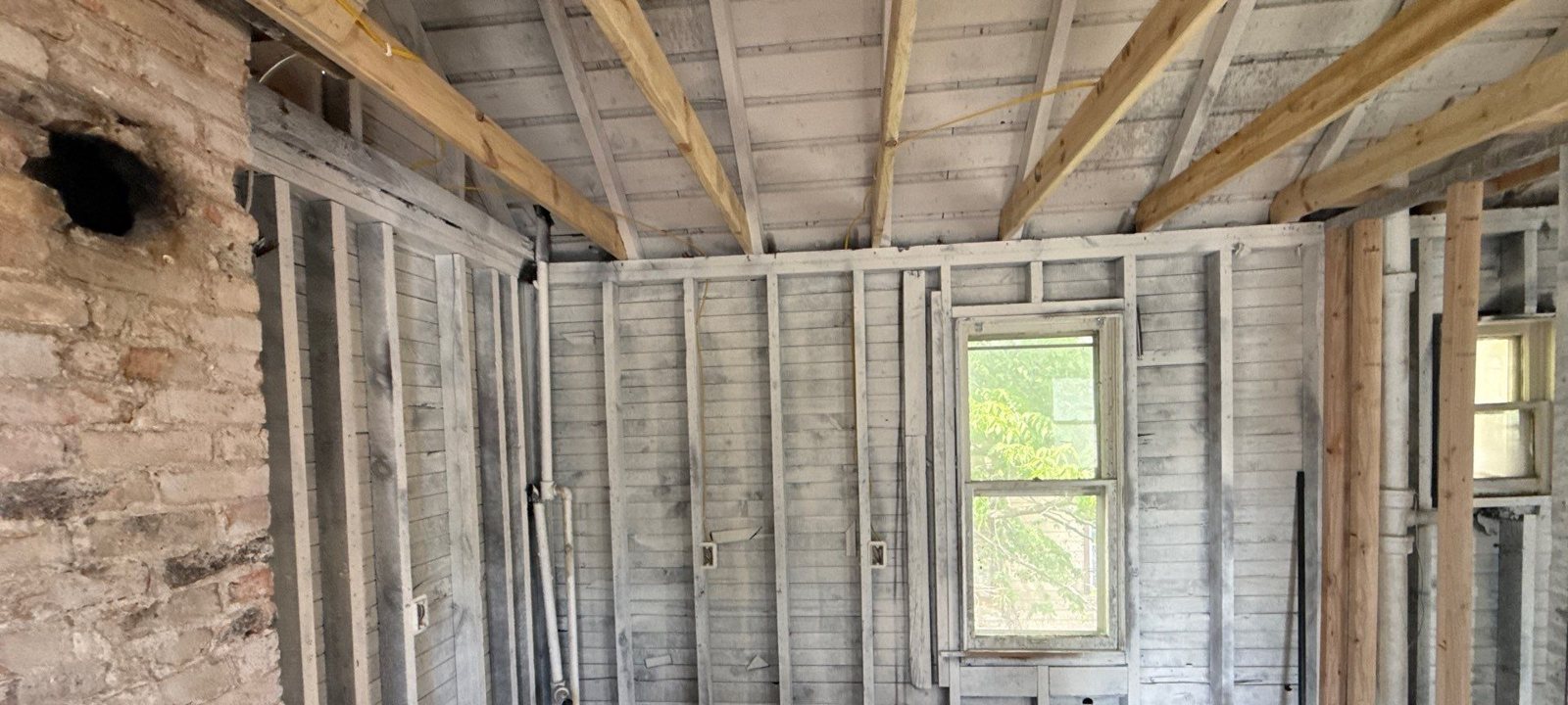 Room with exposed new and original wall studs, ceiling joists, and a window, ready for inspection