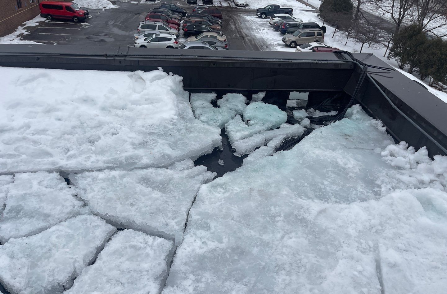 Thick cracked slabs of ice and snow covering a flat roof at the metal roof edge, with standing water between slabs