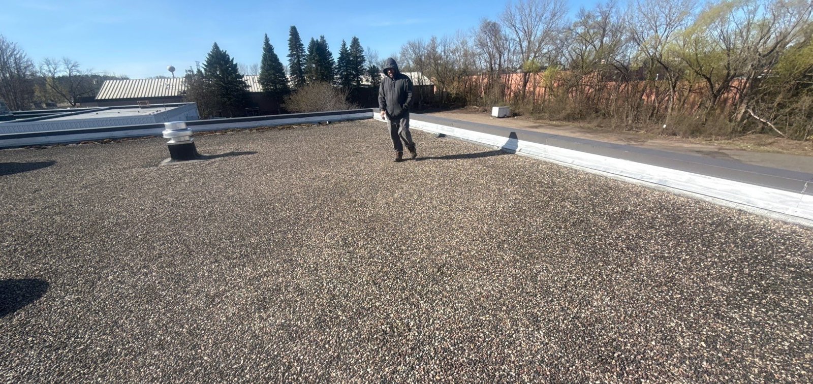 Inspector walking across a gravel-surfaced commercial flat roof with a roof vent and metal edge