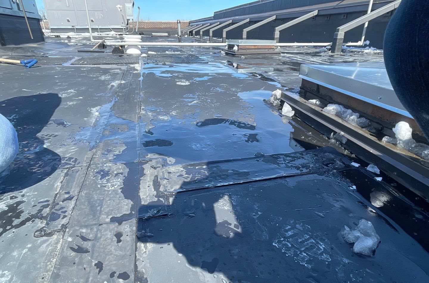 Wet single-ply roof membrane with seams, puddled meltwater, and leftover ice chunks next to a skylight curb
