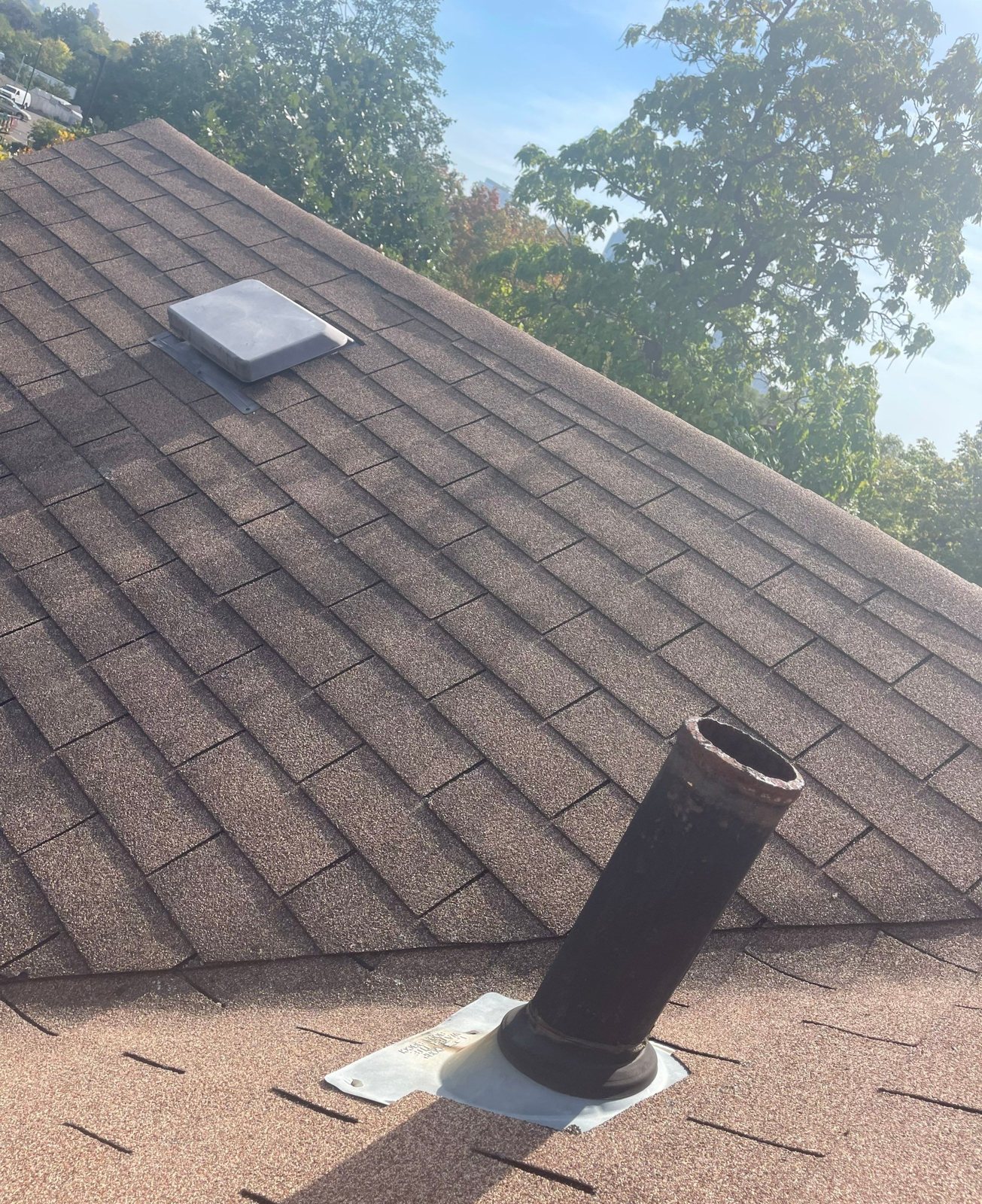 Rusted cast-iron plumbing vent pipe coming through a brown asphalt shingle roof, with a box vent behind it