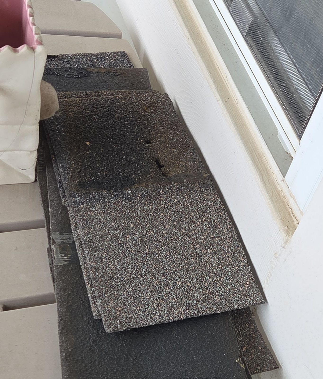 A stack of torn asphalt shingle pieces that blew off a roof, set on a deck beside the house