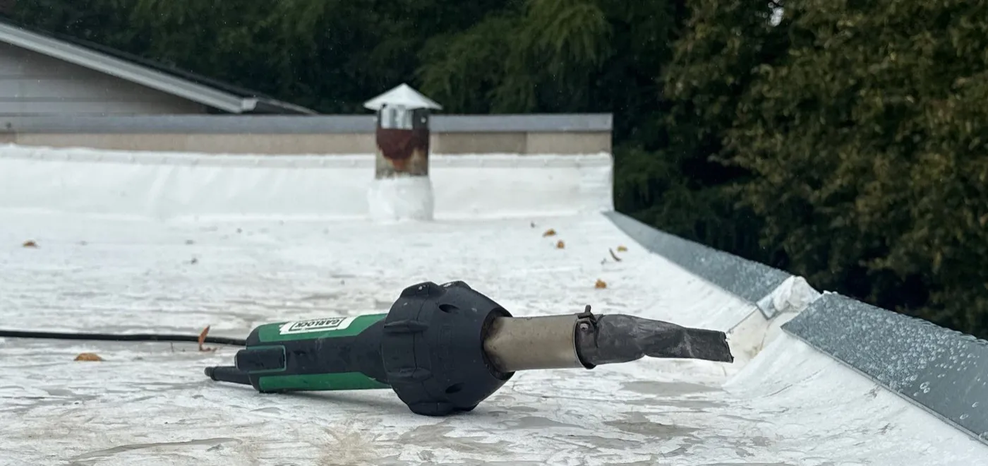 Hot-air hand welder resting on a white thermoplastic flat roof membrane next to a parapet with metal coping
