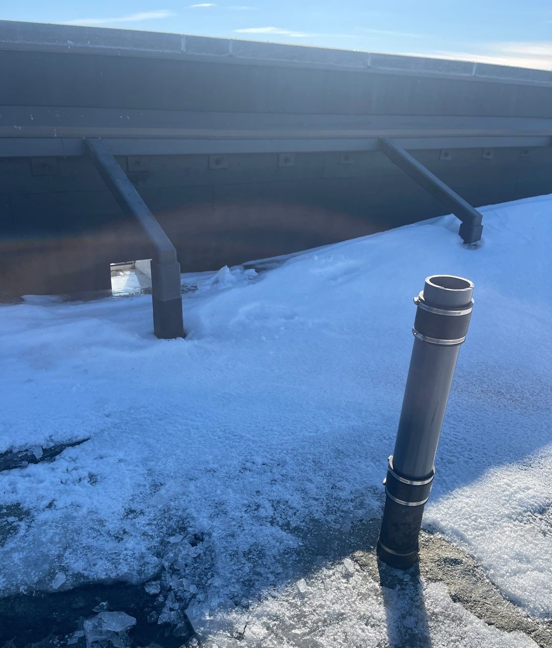 Vent pipe sticking up through packed snow on a roof below a gutter line