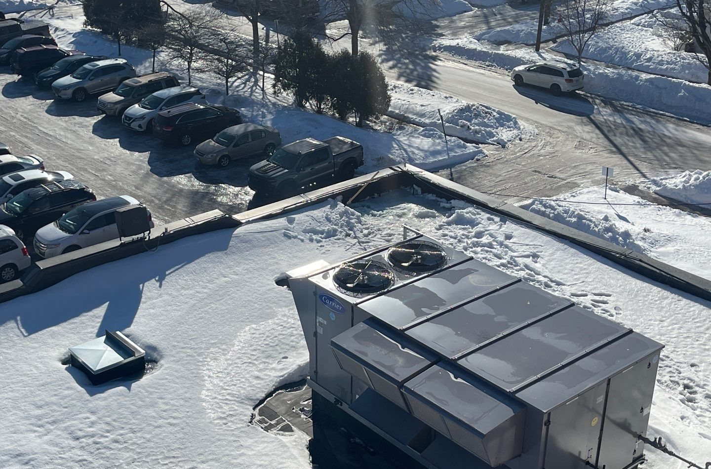 Snow and ice covering a flat rubber commercial roof with a rooftop unit, parking lot below