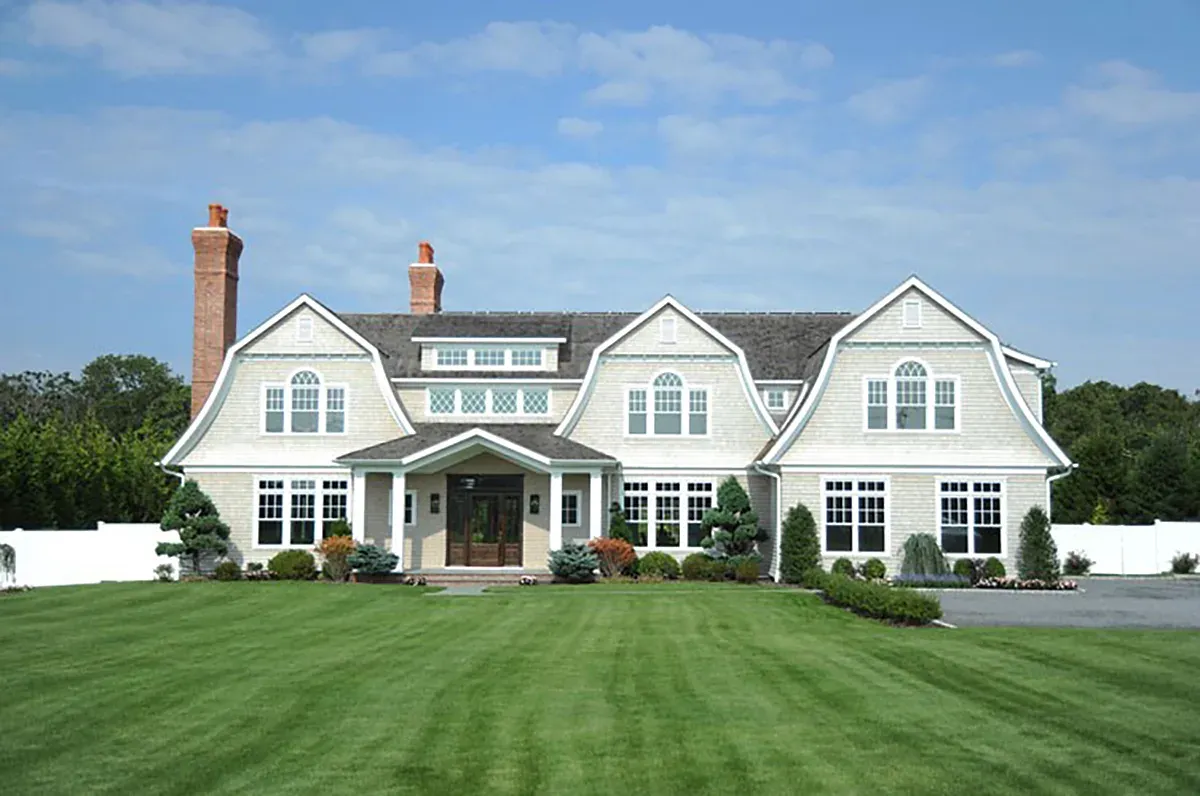 A shingle-style Prestige home with a gambrel roof, white trim and two chimneys, on a wide green lawn under a clear sky