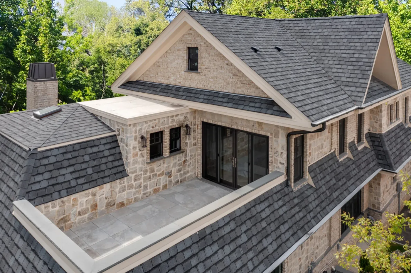 The rear of a stone and shingle Prestige home, a balcony above the wide dark-framed glass doors