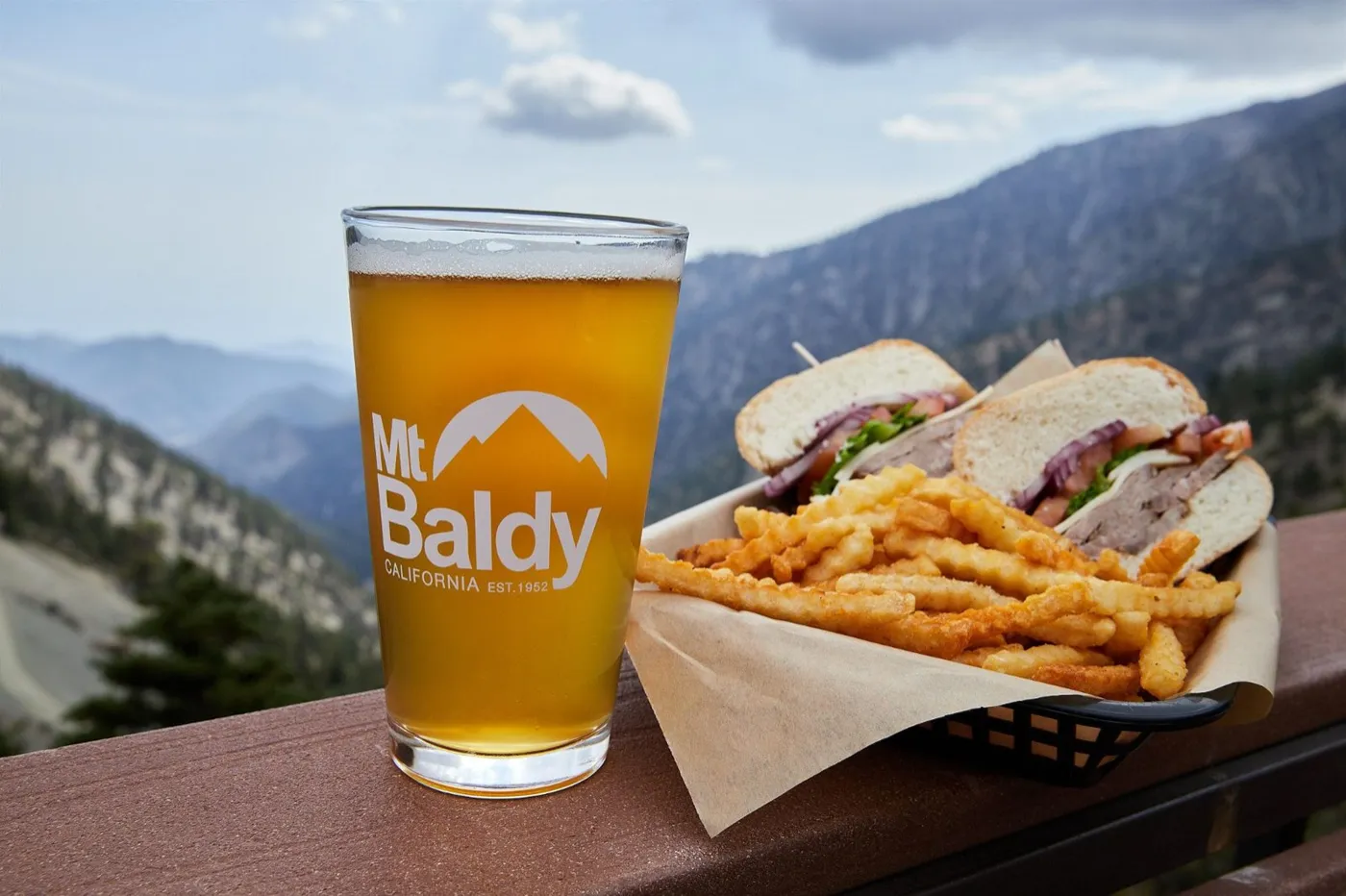 A golden pint in a Mt Baldy Brewery glass beside a basket of fries and a sandwich, with mountains behind