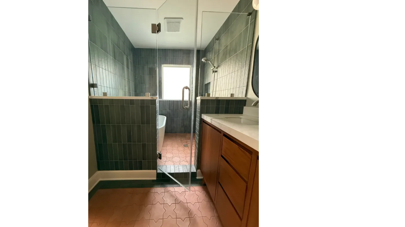 Walk-in glass shower in a sage green tile bathroom with terracotta hex floor and wood vanity
