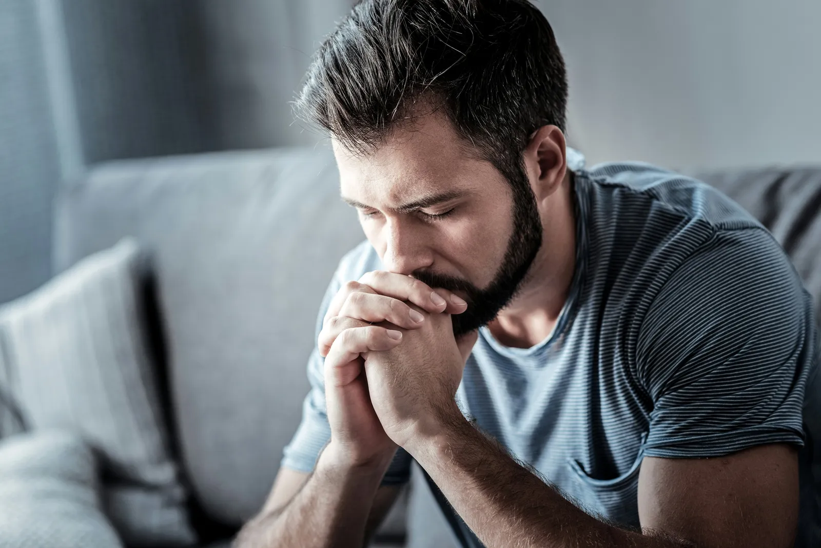 A man sitting on a couch, eyes closed, hands clasped in front of his face