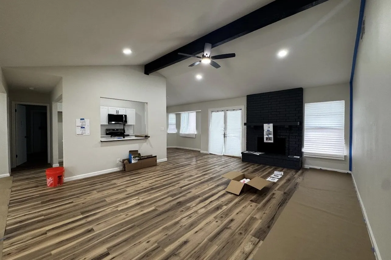 Living room mid-build: vaulted ceiling with a dark beam, painted brick fireplace, new floors, boxes on the floor