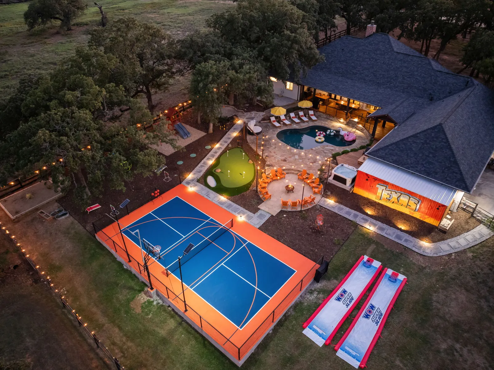 Whole backyard at dusk from above: multi-use court, putting green, pool, fire pit circle and covered patio