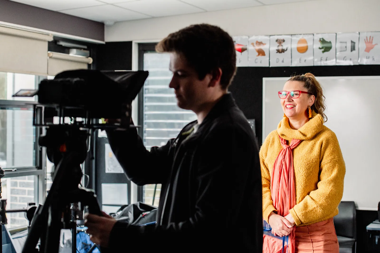 Camera operator framing a classroom interview with a woman in a yellow cardigan