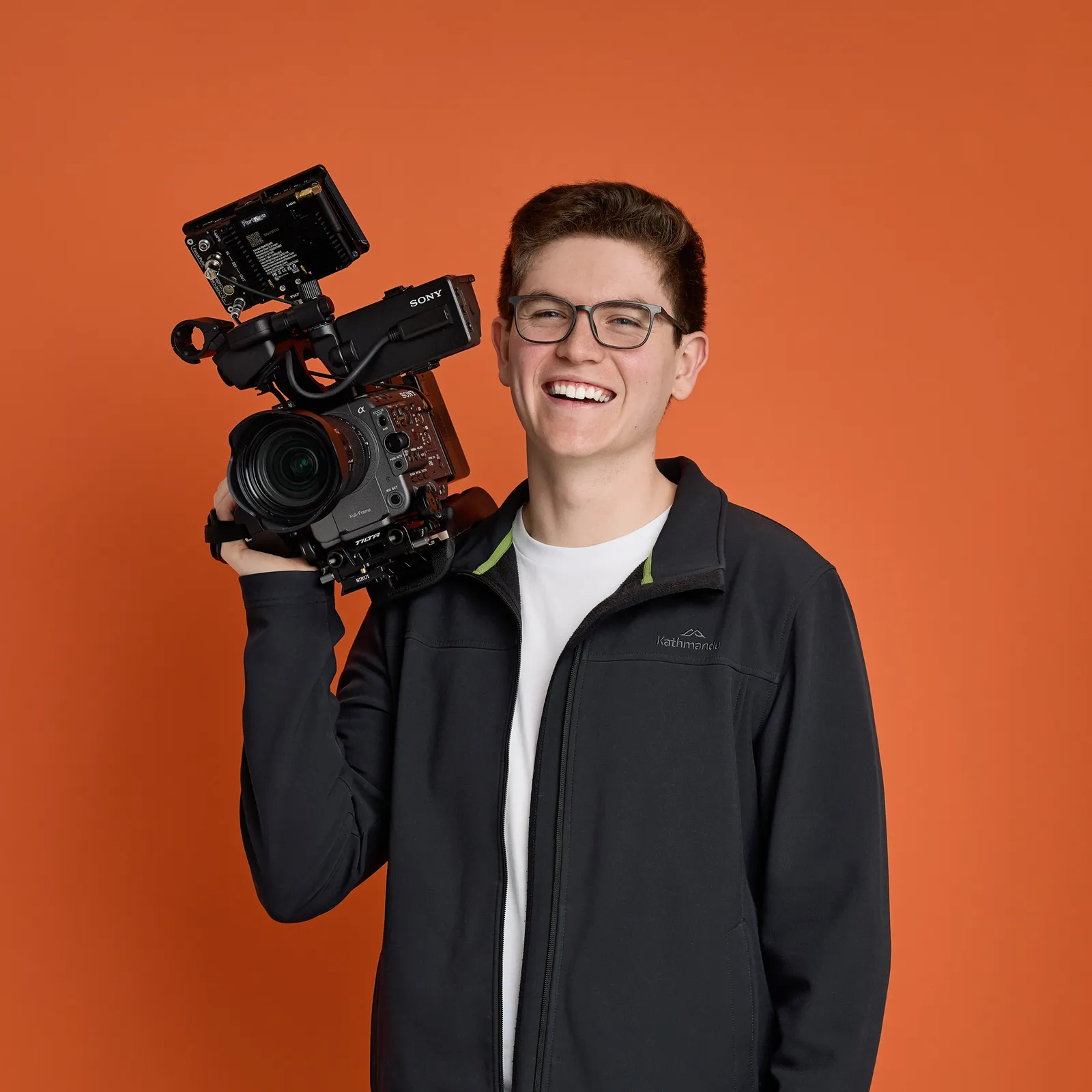 Henry Sheahan smiling with a cinema camera on his shoulder against an orange backdrop