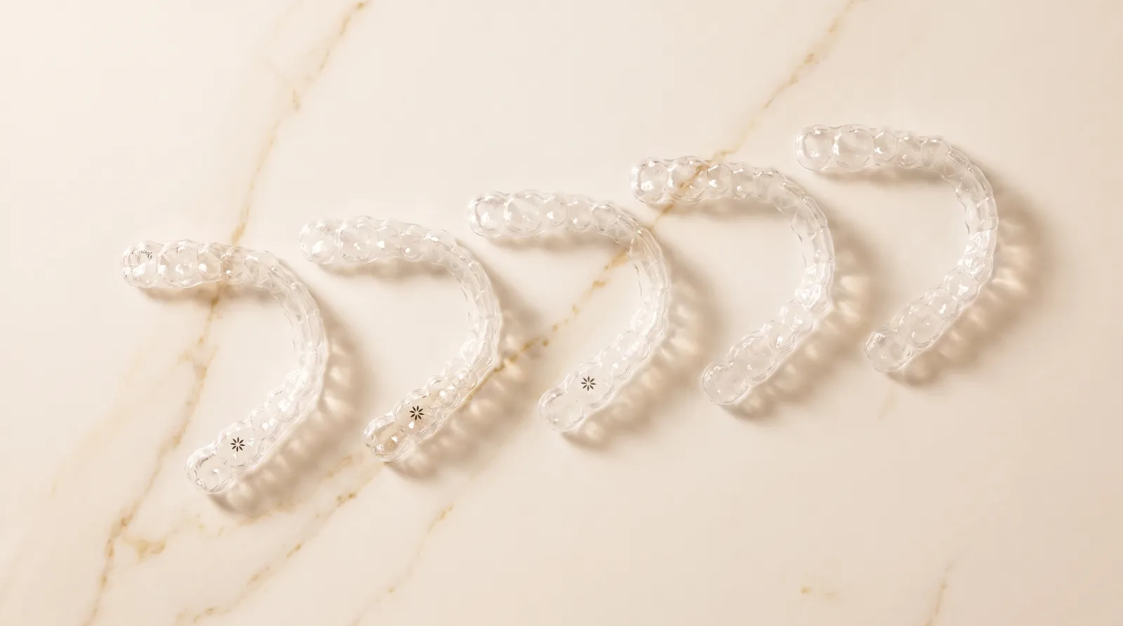 Flatlay editorial: cinco alineadores Invisalign transparentes en progresión diagonal sobre mármol cálido — cada uno una etapa del plan de tratamiento