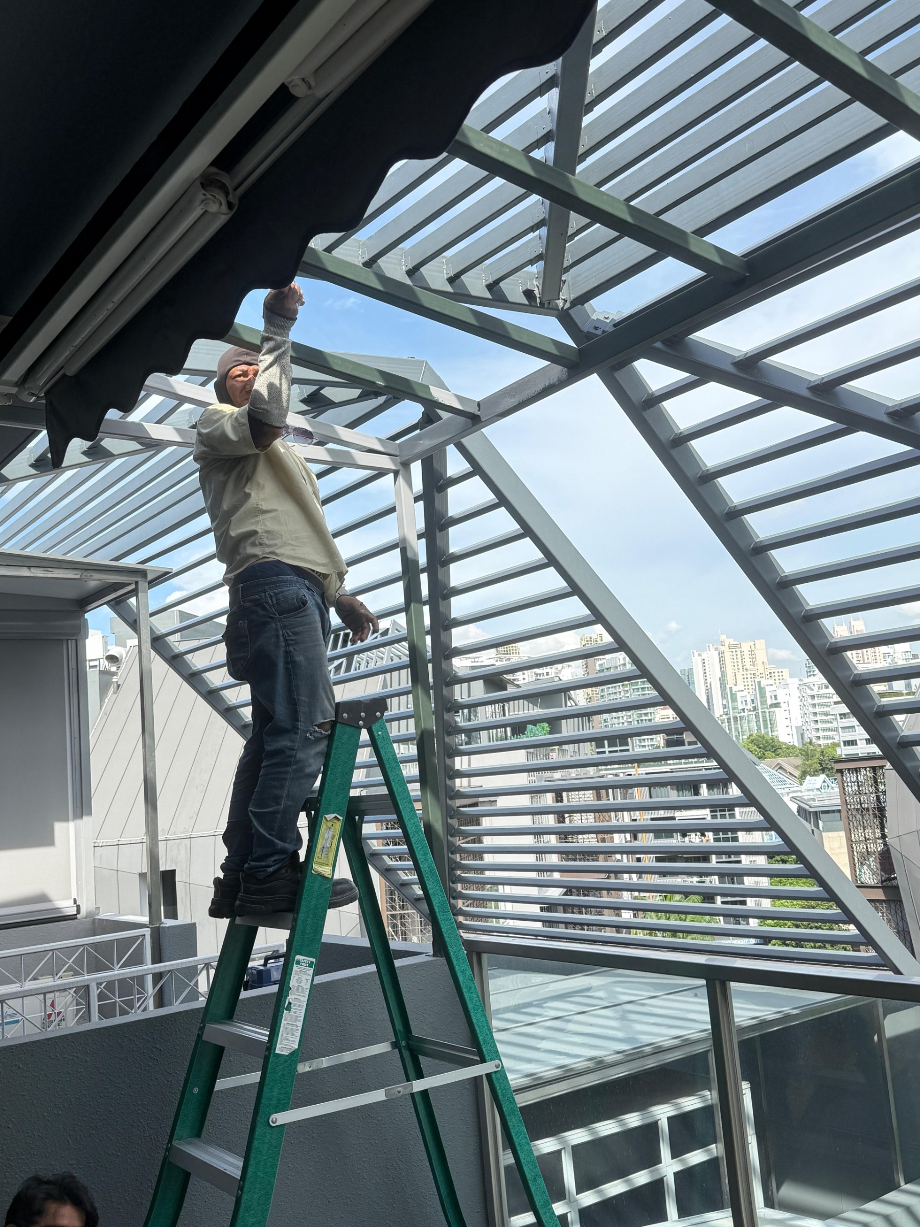 Installer on a ladder fitting a shade canopy under a louvre roof frame