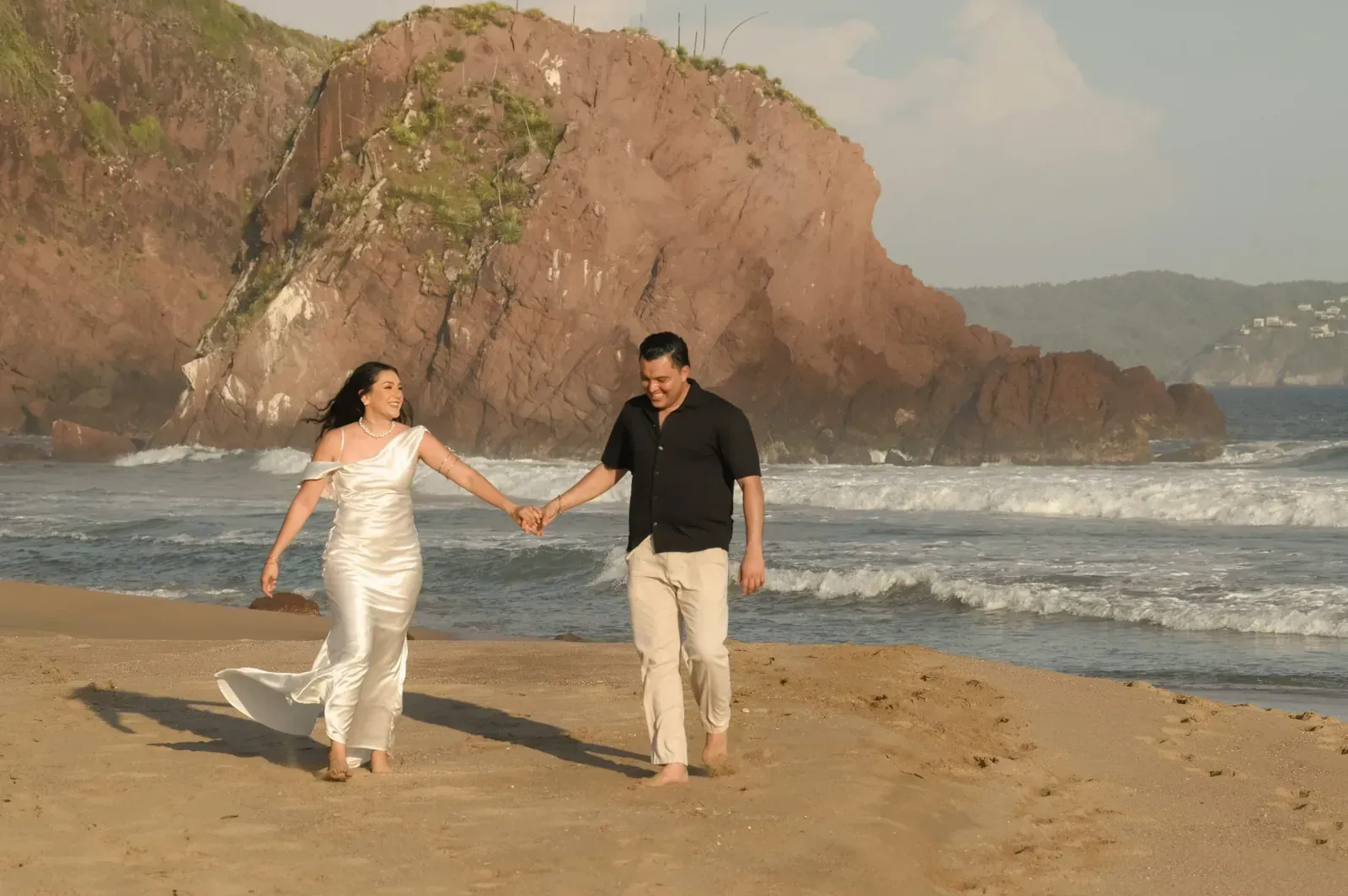 Isamar y Brian caminan de la mano hacia el mar, junto a una gran roca