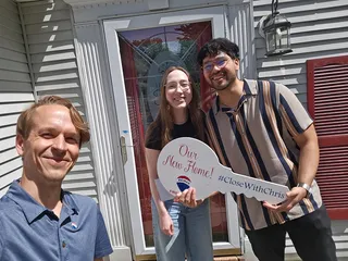 Three people with a large home key sign by a front door.