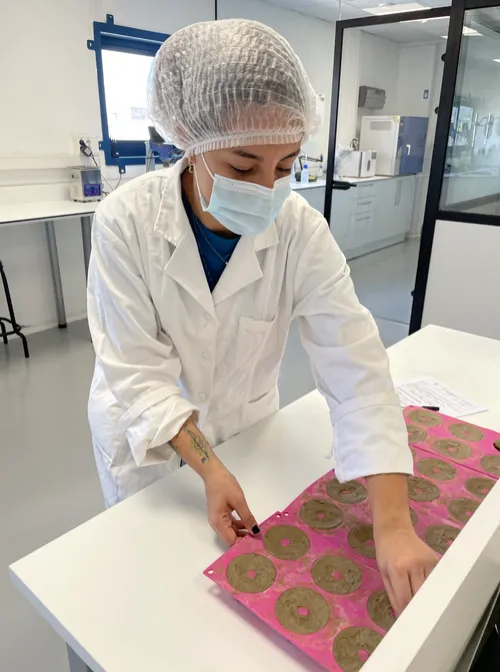 Lesly en un laboratorio, con bata, gorro y mascarilla, trabajando sobre una mesa
