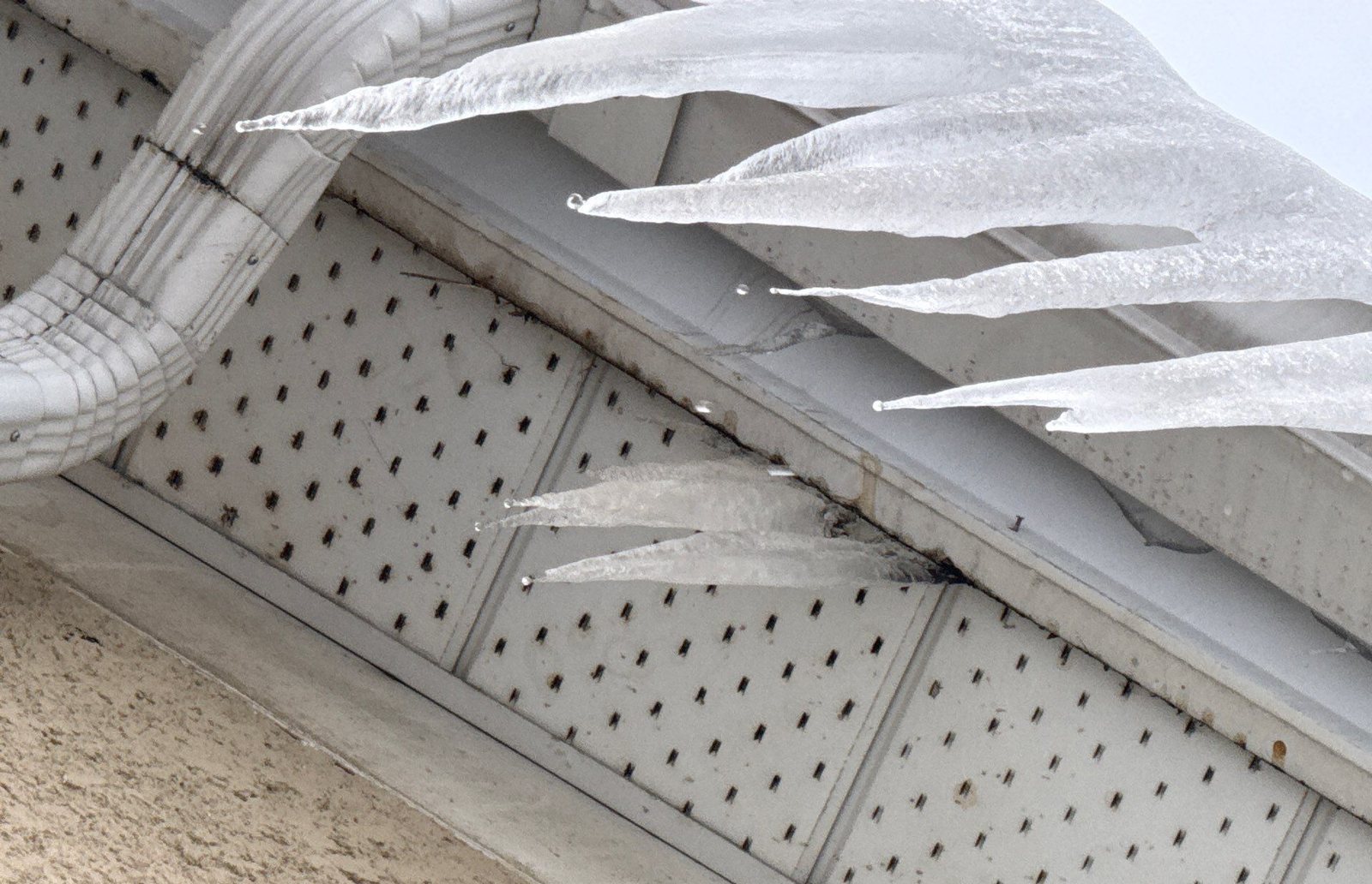 Long icicles hanging from a roof edge and behind the gutter above a vented soffit and downspout