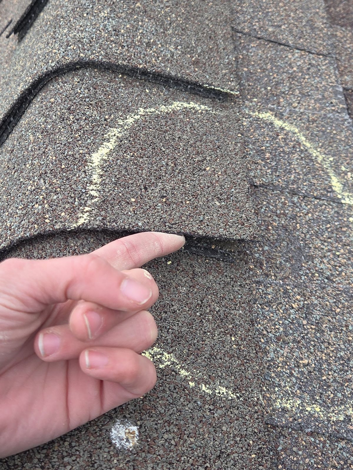 Inspector's finger pointing under a lifted asphalt shingle tab that has come unsealed, with the damaged area circled in yellow chalk