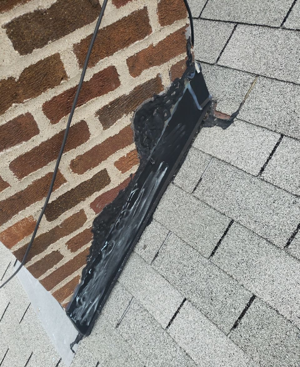 Brick chimney on a gray shingle roof with its flashing covered in thick black tar patching