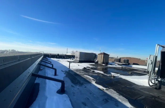 Flat commercial roof with snow packed along the parapet wall, partly cleared membrane, and rooftop units in the background