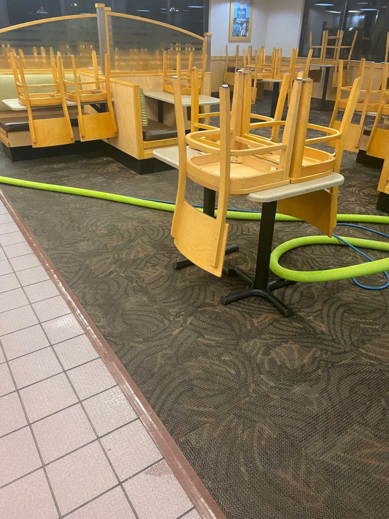 Restaurant dining room with chairs stacked on yellow tables and a hose across the patterned carpet during cleaning