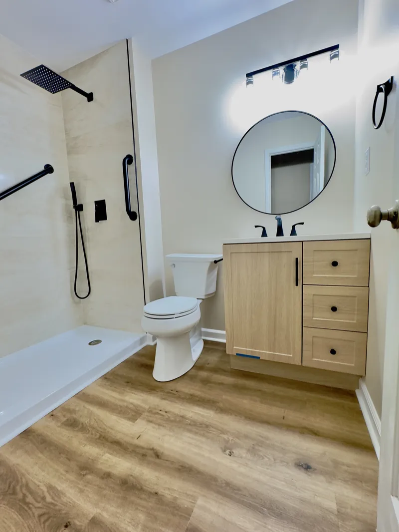 Compact bathroom with beige shower walls, black fixtures, light wood vanity and wood-look floor