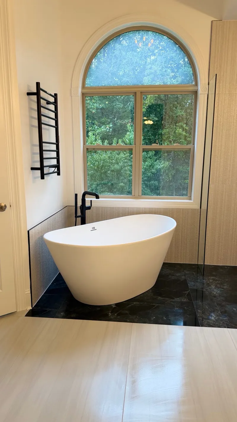 Freestanding white bathtub beneath an arched window with black fixtures and a towel warmer