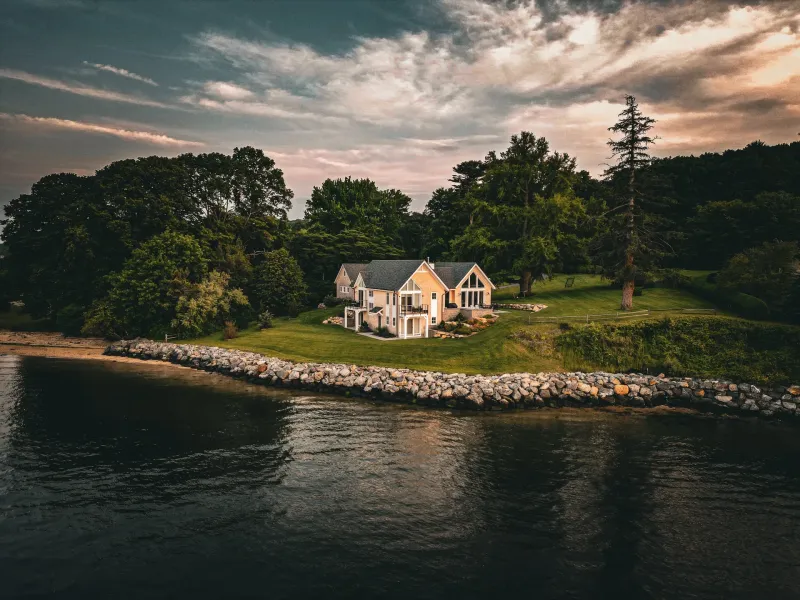 East Hills Renovation: a shingled waterfront house with tall gable windows above a lawn and stone seawall at dusk