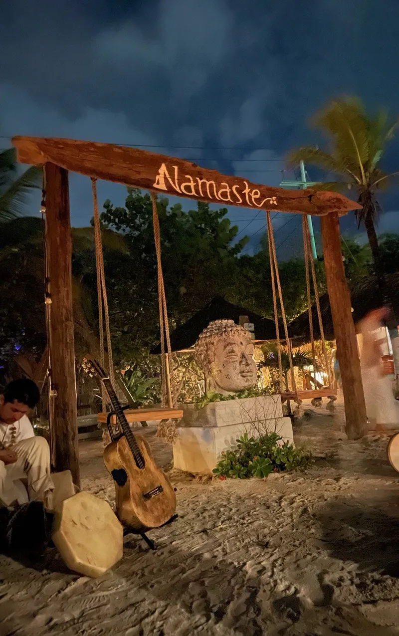 Cartel de Namasté sobre la arena, con columpios y palmeras