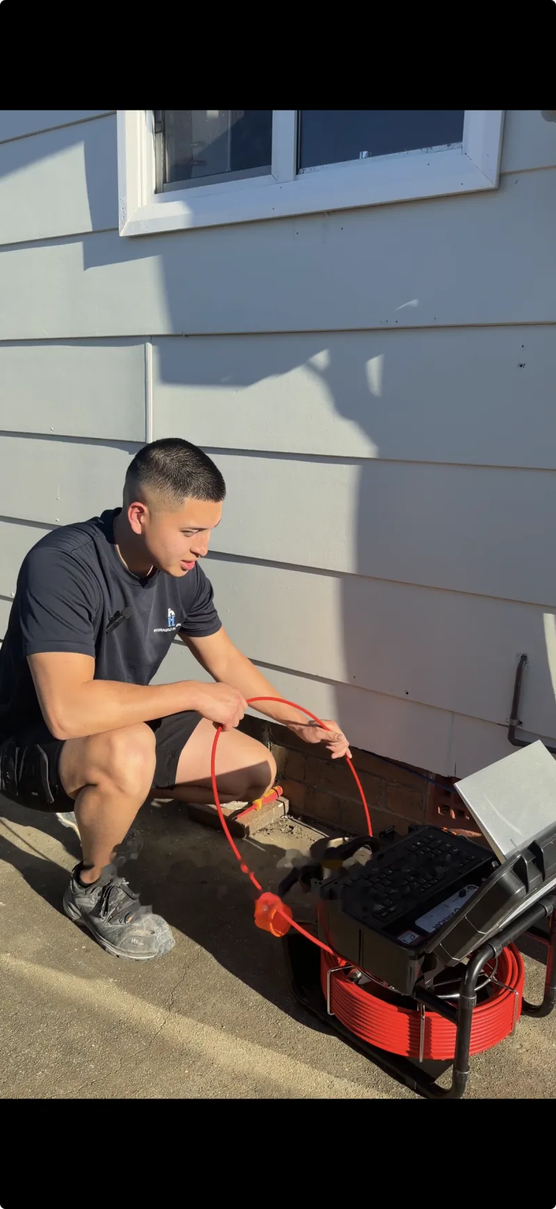 Plumber feeding a CCTV drain camera on a reel into a drain access point beside a house