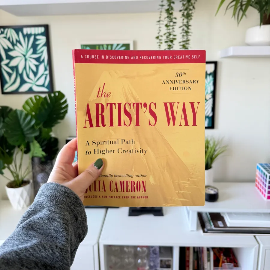 A hand holding up the yellow Artist's Way book in a bright art studio with plants and framed prints.