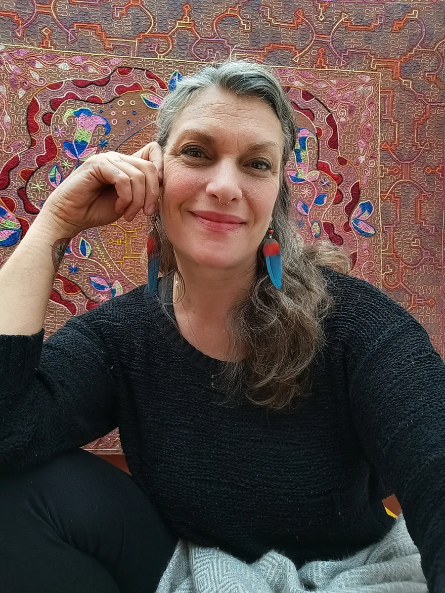 Jessa Frymire smiling, wearing feather earrings, in front of a Shipibo embroidered textile