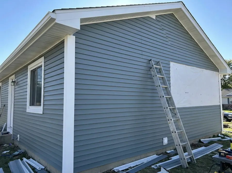 Blue-grey vinyl siding being installed with white soffit and fascia, house wrap still showing on one section