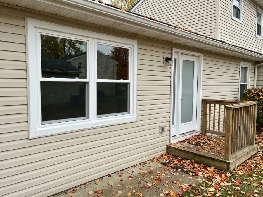 New white double-hung windows and a new entry door in a beige vinyl-sided wall, autumn leaves on the ground